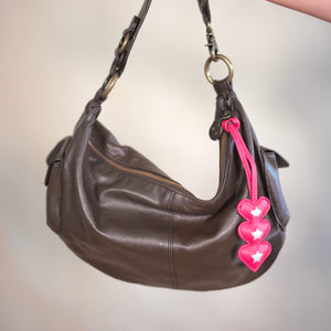Brown leather handbag with pink heart-shaped tags held by a hand against a neutral background